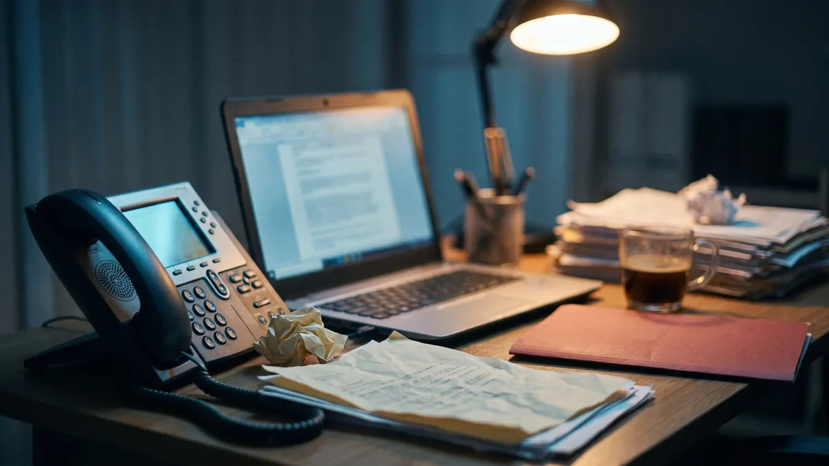 Teléfono de escritorio y computadora encendida de noche con papeles desordenados