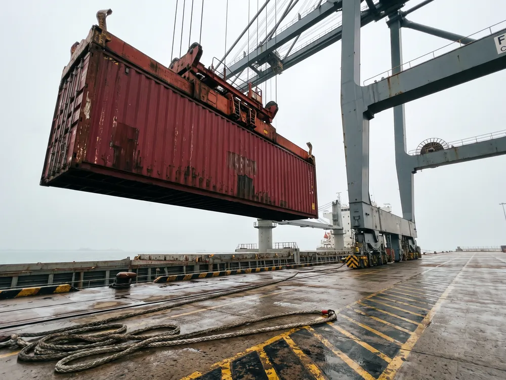Contenedor rojo suspendido de una grúa de muelle mientras baja hacia el buque